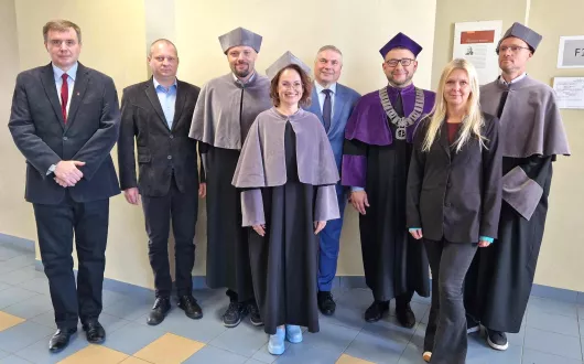 Kolegium dziekańskie Wydziału FTIMS w togach dziekańskich oraz Dyrektorzy Instytutów w eleganckich strojach na tle beżowej ściany w budynku B9.