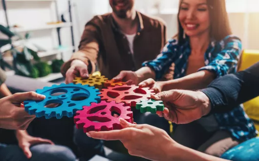 Grupa młodych osób w kolorowych ubraniach łączy duże, piankowe, kolorowe puzzle w kształcie kół zębatych.