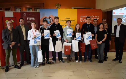 Młodzi laureaci konkursu prezentują dyplomy w towarzystwie wykładowców z PŁ. W tle roll-upy Wydziału FTIMS PŁ oraz Zespołu Szkół Nr 2 w Wieluniu 