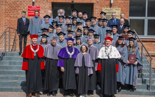 Uczestnicy rozdania dyplomów w togach i biretach pozują do pamiątkowego zdjęcia na schodach gmachu Instytutu Fizyki