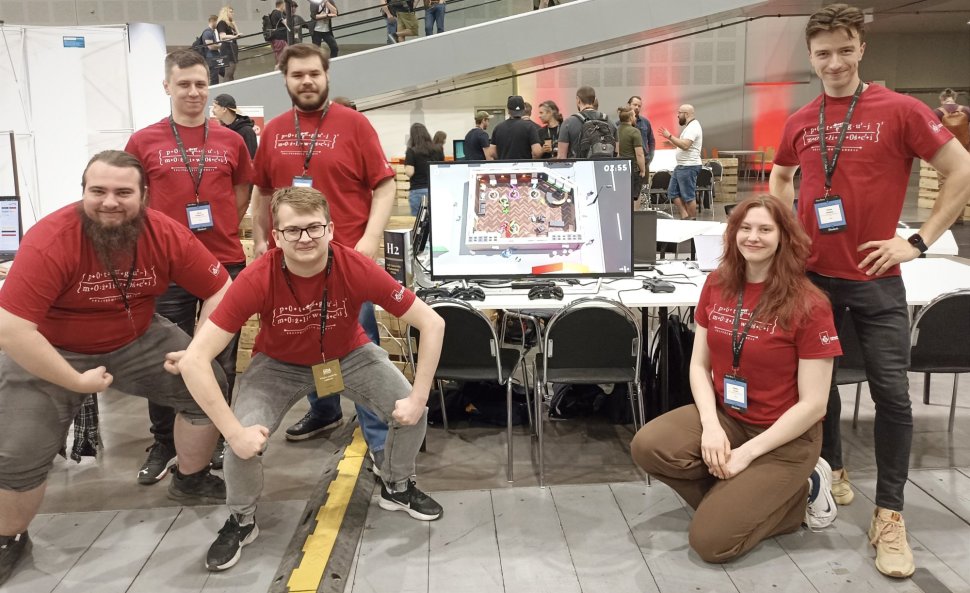 Sześcioro uśmiechniętych studentów w czerwonych koszulkach PŁ pozuje do zdjęcia przy stanowisku komputerowym. Pomiędzy nimi duży monitor ukazujący kadr z gry komputerowej