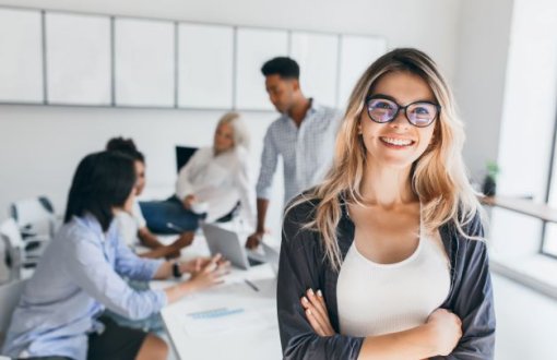 Uśmiechnieta blondynka w okularach ze skrzyżowanymi ramionami. W tle grupa studentów pracujących nad projektem